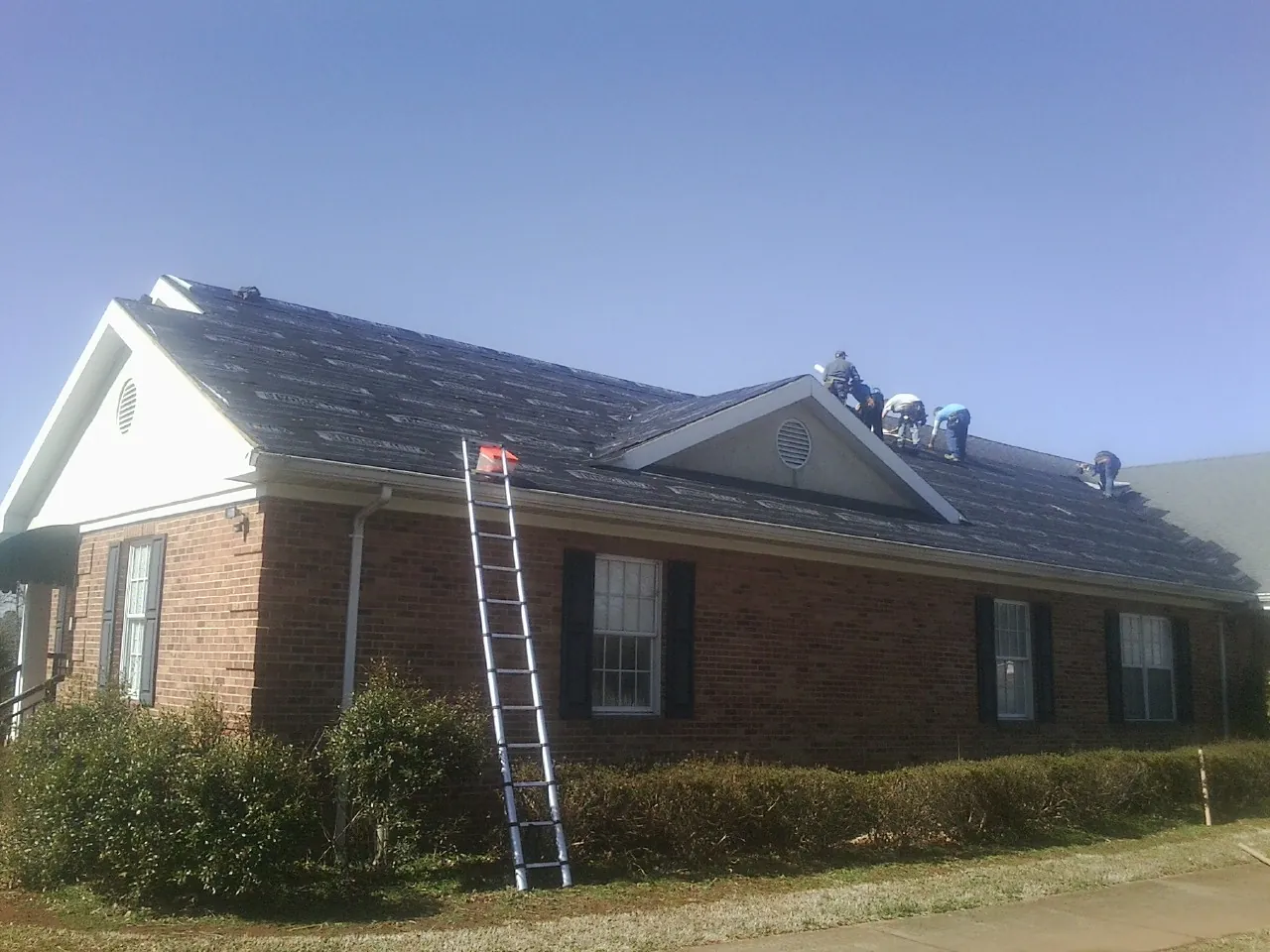 Crew tearing shingles off a brick ranch roof with a ladder set — Emergency Roof Repair work by our crew in Nora Springs