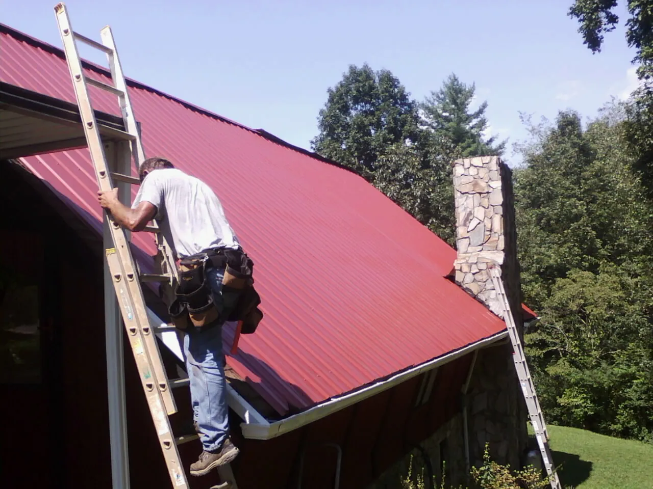 Roofer on a ladder beside a red metal roof and stone chimney — Soffit & Fascia Repair by Roofing Brothers in Nora Springs